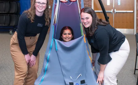 A child sits inside a suspended tent and two students pose around her