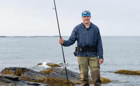 A UNE student poses with a fishing rod in front of the ocean