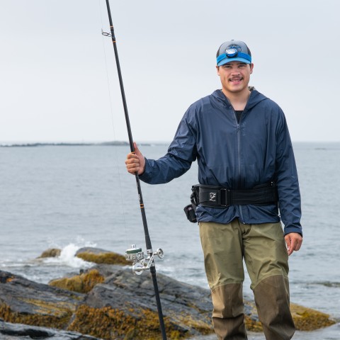 A UNE student poses with a fishing rod in front of the ocean