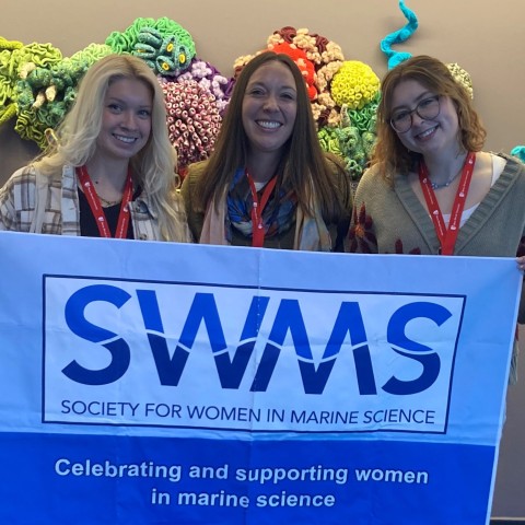 Three UNE researchers pose holding a Society for Women in Marine Science banner